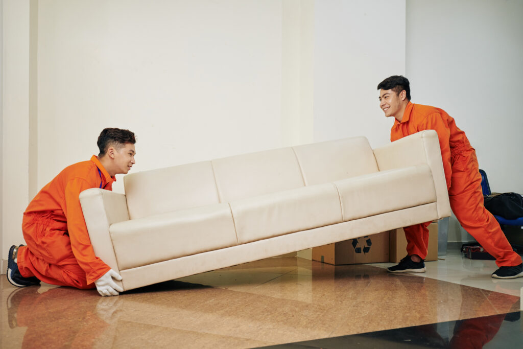 Smiling moving service company workers in uniform carrying heavy couch